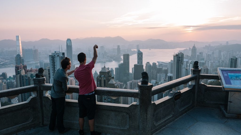 Photo by Robson Hatsukami Morgan two men standing on top of building pointing to city at daytime